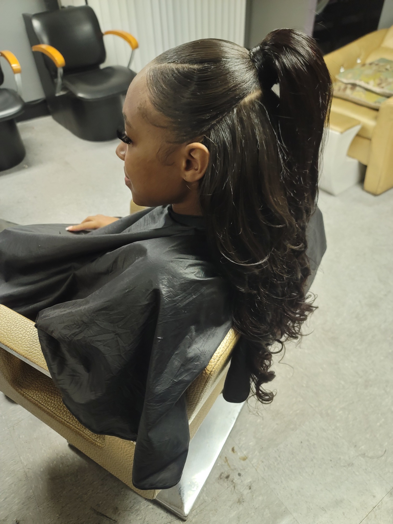 Girl with long, wavy hair styled in a high ponytail, seated in a salon chair.